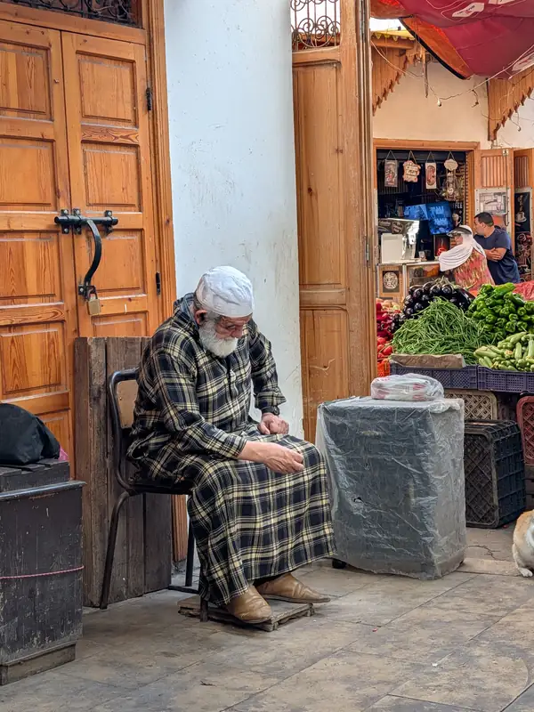 Un marchand âgé assis devant une porte de boutique, occupé à ses mains, des légumes sur l'étal voisin